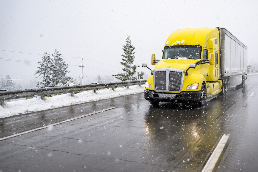 Yellow semi truck drives on highway through snow